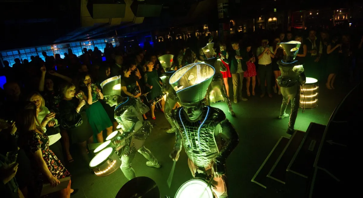 Spark! performers drumming through a crowded party in green-tinted light, guests dancing.