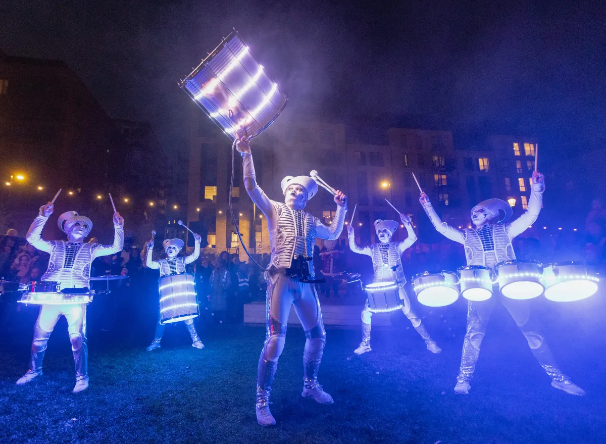 Spark! performers drumming at night in a park, one lifting an illuminated drum overhead, lit purple and white.