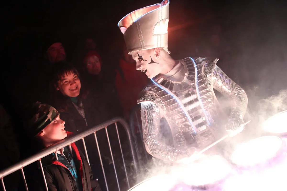 A Spark! performer leaning over a barrier to play to two delighted children, drums glowing.