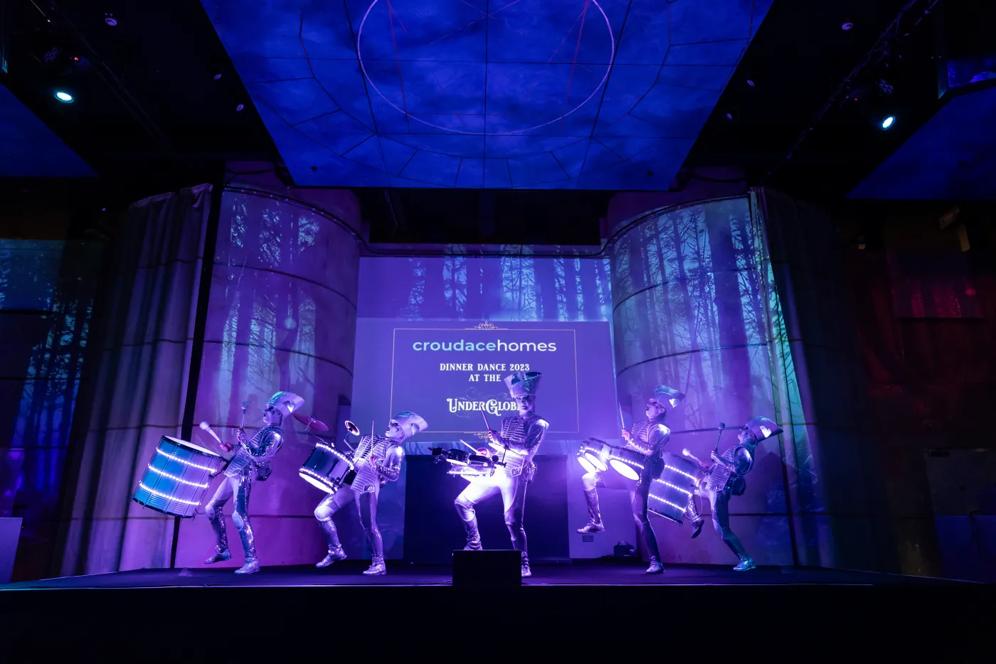 The Spark! cast on the stage of a company dinner dance, one performer lifting a glowing drum.