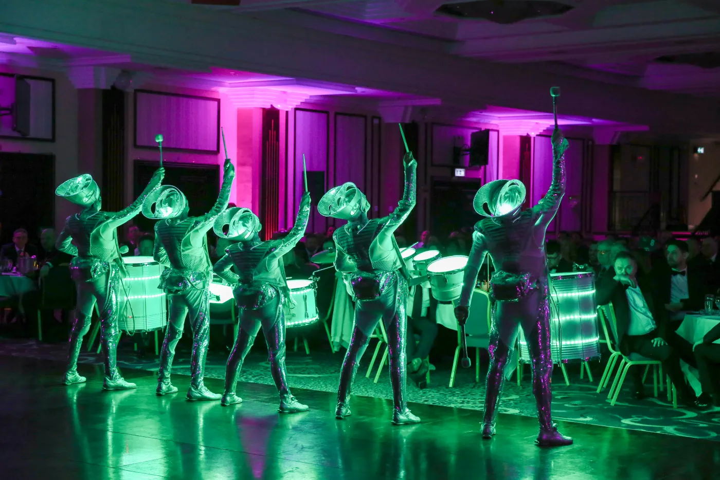Five Spark! performers lit green, drumming beside the dinner tables in a hotel ballroom.