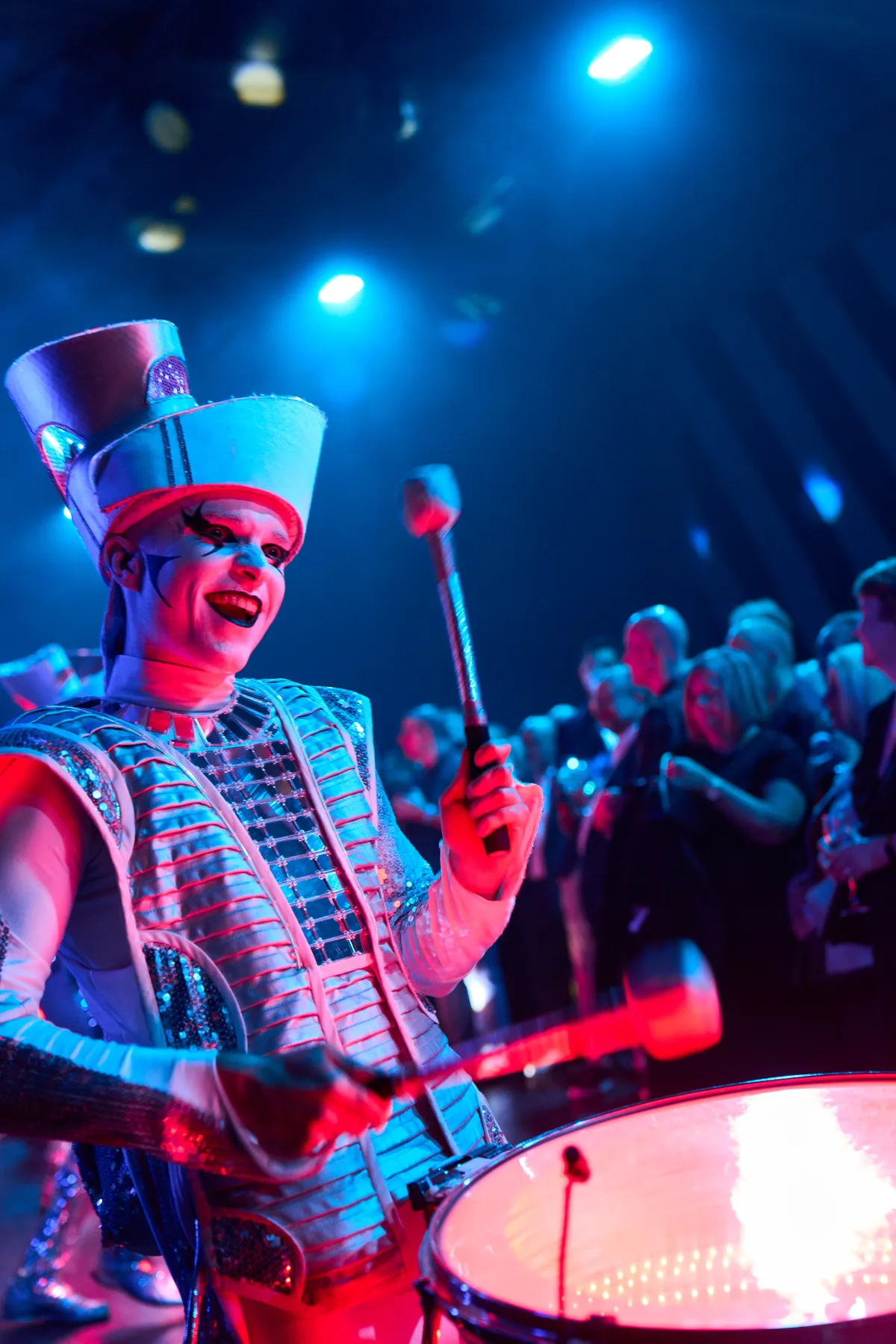 A Spark! performer grinning mid-beat, sticks raised, lit red and blue.