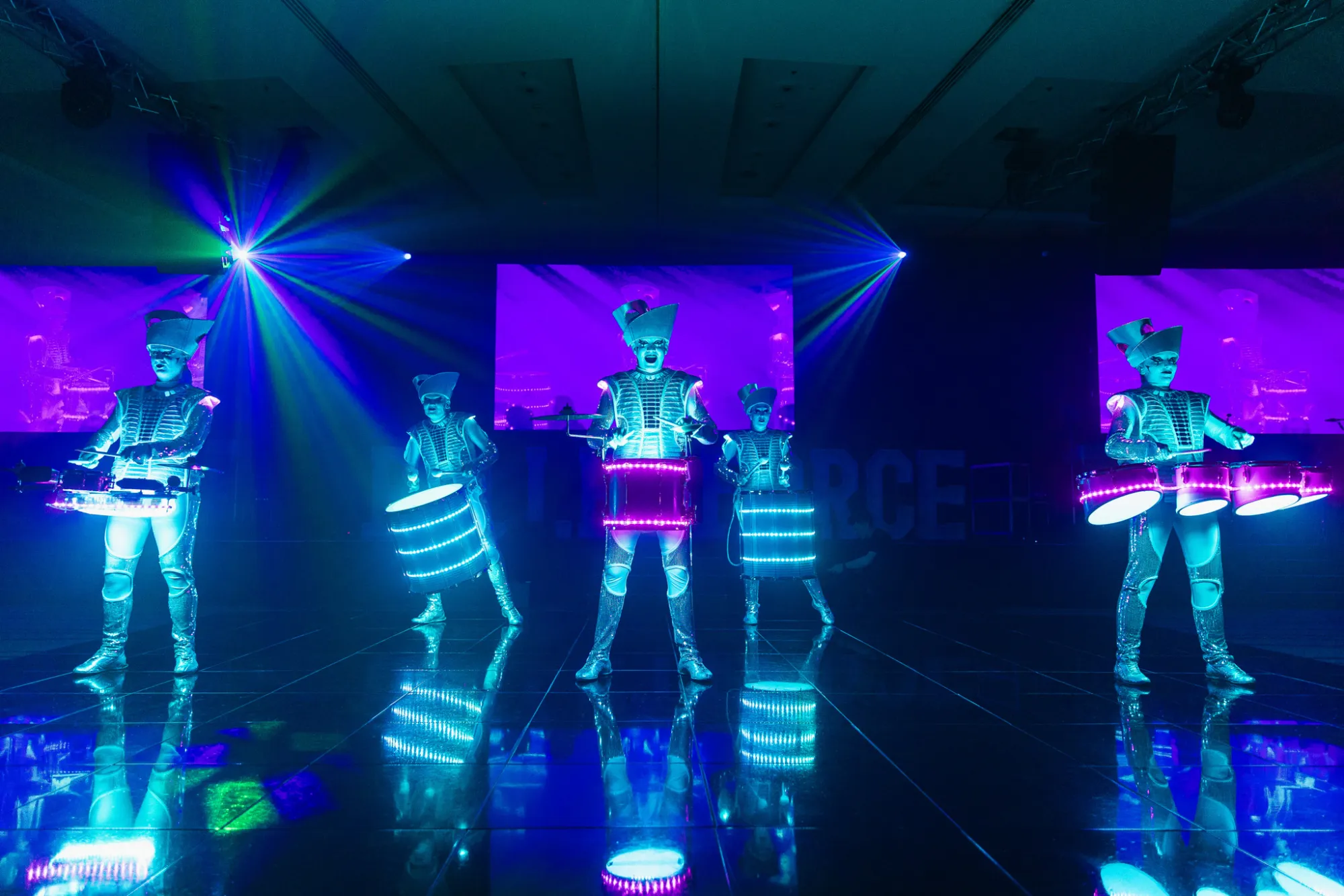 Three Spark! performers drumming on a mirrored ballroom floor, lit electric blue, screens glowing behind them.