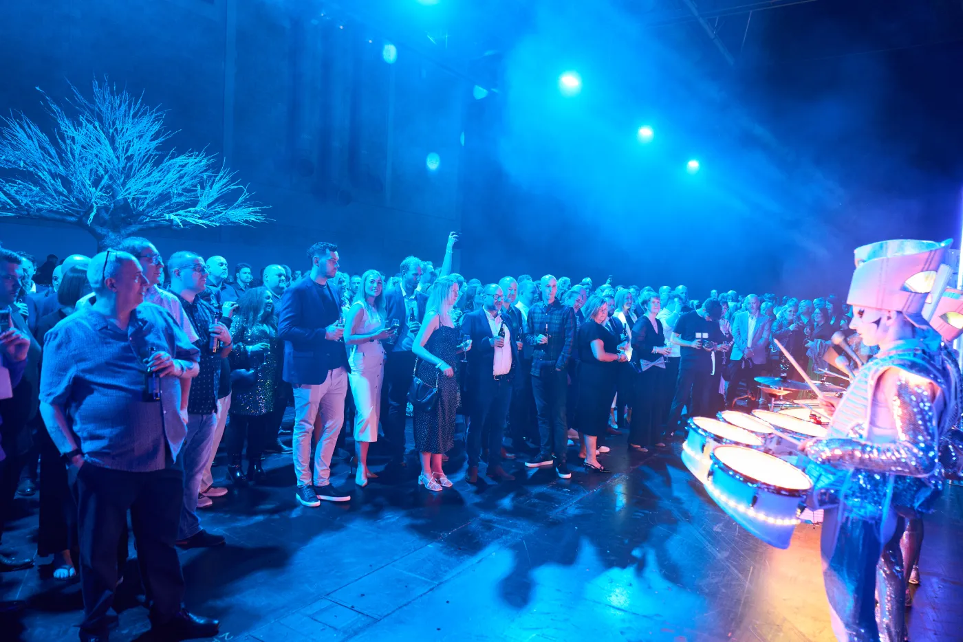 A Spark! performer drumming beside a standing crowd at a company party, blue light and haze.