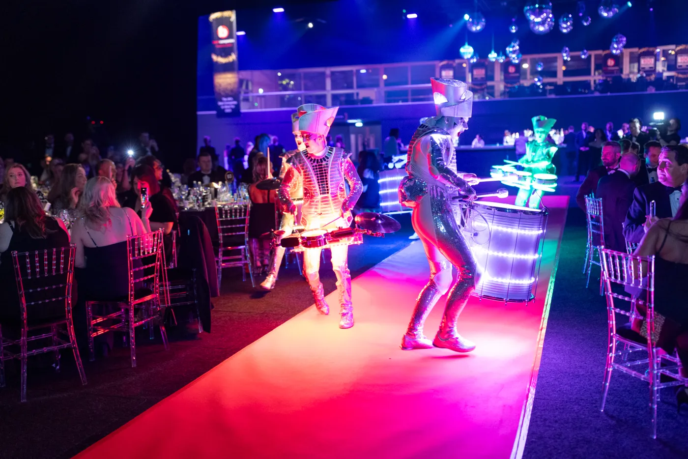 Spark! performers drumming down a runway between dinner tables, lit magenta, guests turning in their seats.