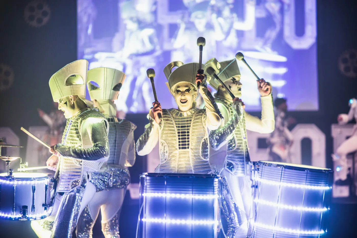 Three Spark! performers close up in a ballroom, masks and illuminated drums glowing gold and white.