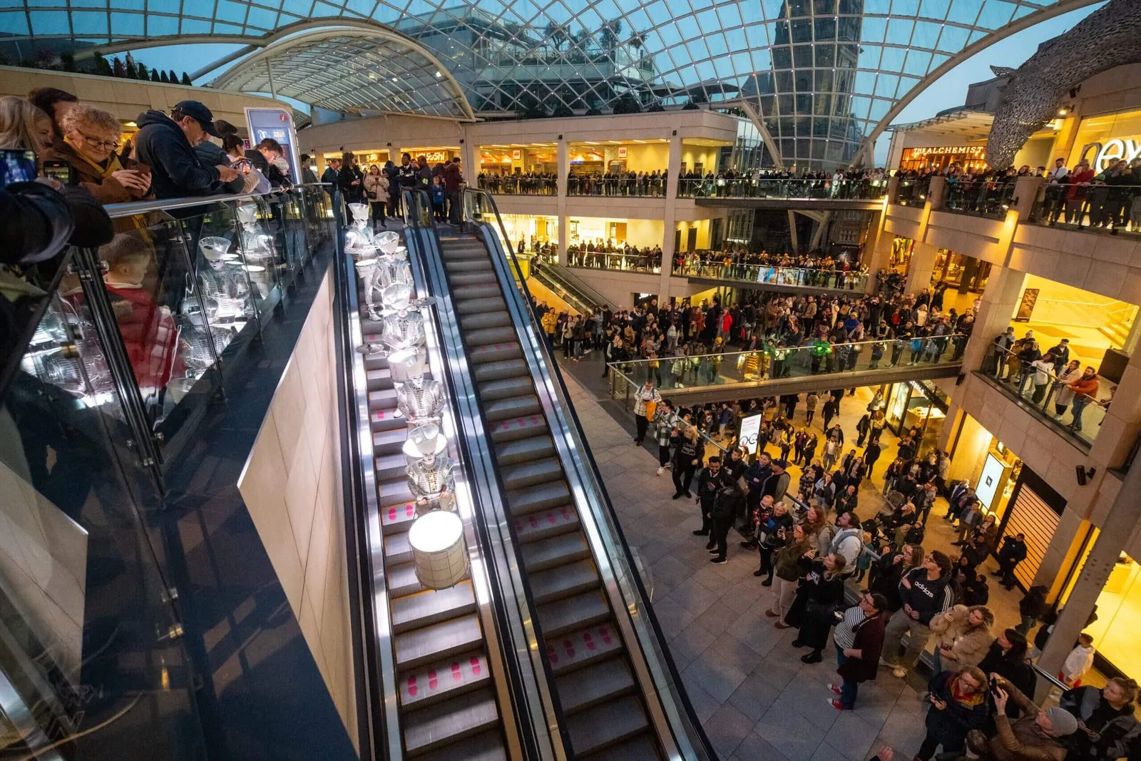 Spark! performers descending an escalator in a shopping centre as a huge crowd watches from every balcony.