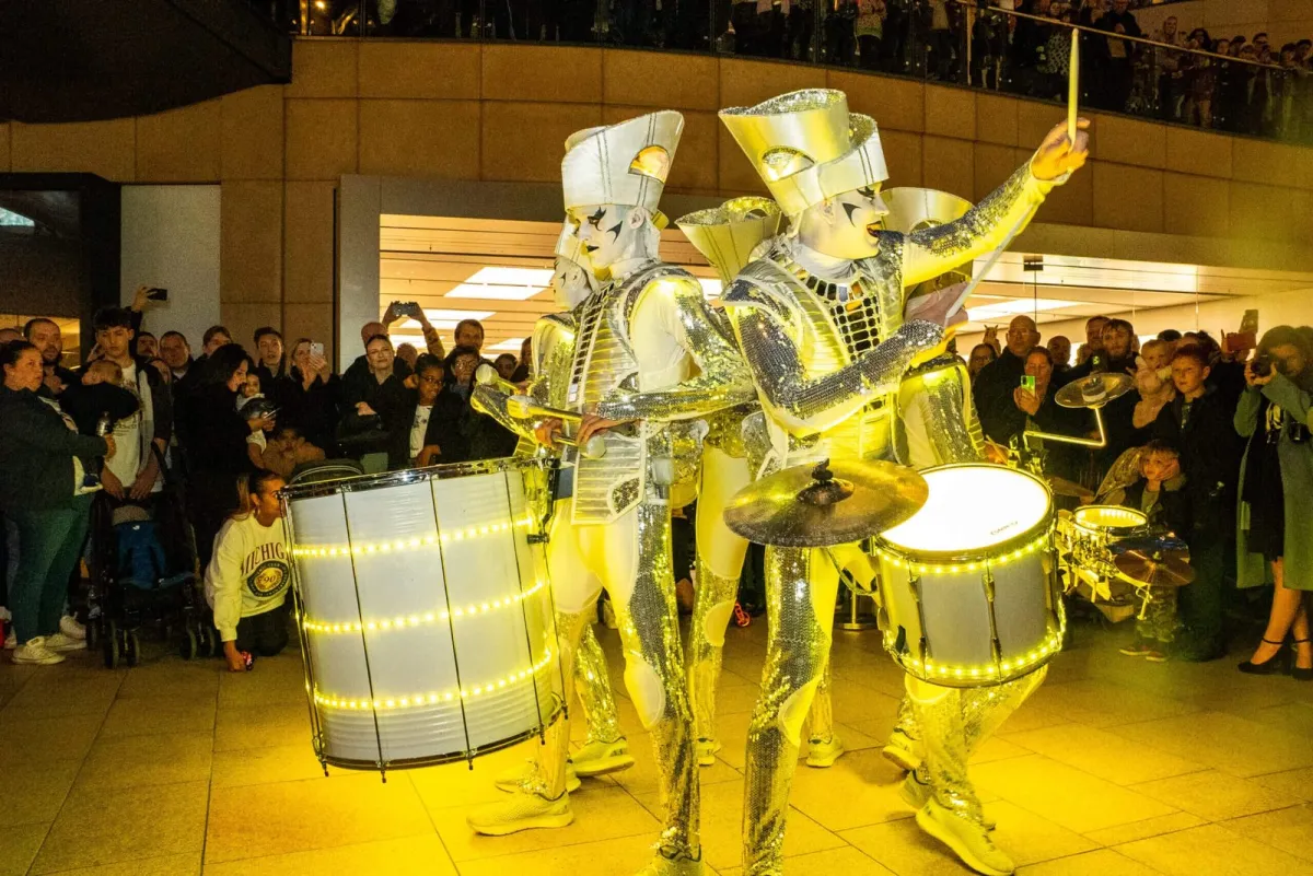 Spark! performers in mirrored gold costumes drumming in a shopping centre as shoppers watch from the balconies.