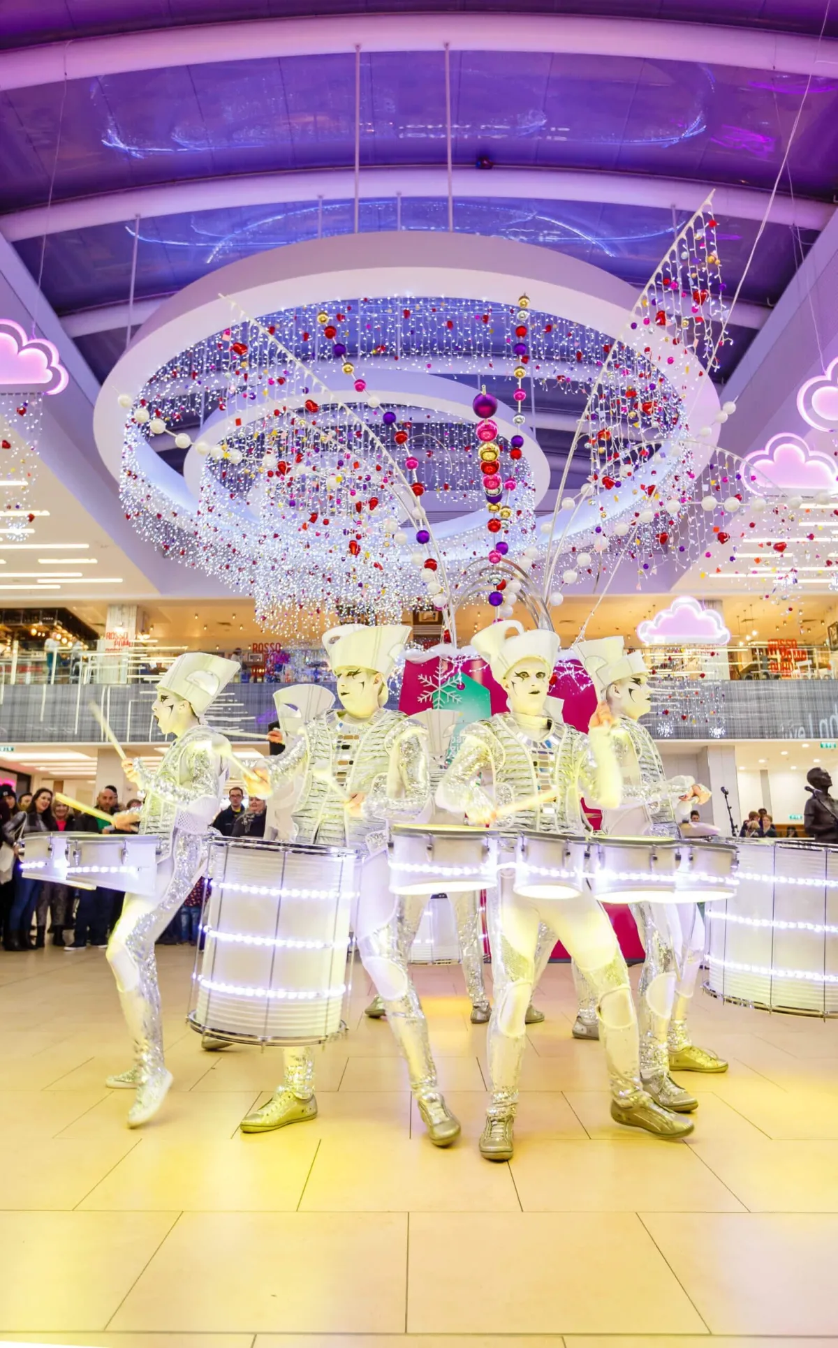 Spark! performers in white and gold drumming beneath giant Christmas decorations in a shopping centre.