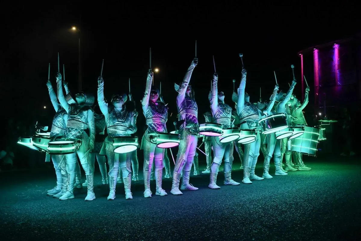 A line of Fireflies performers with drumsticks raised together, lit teal and green.