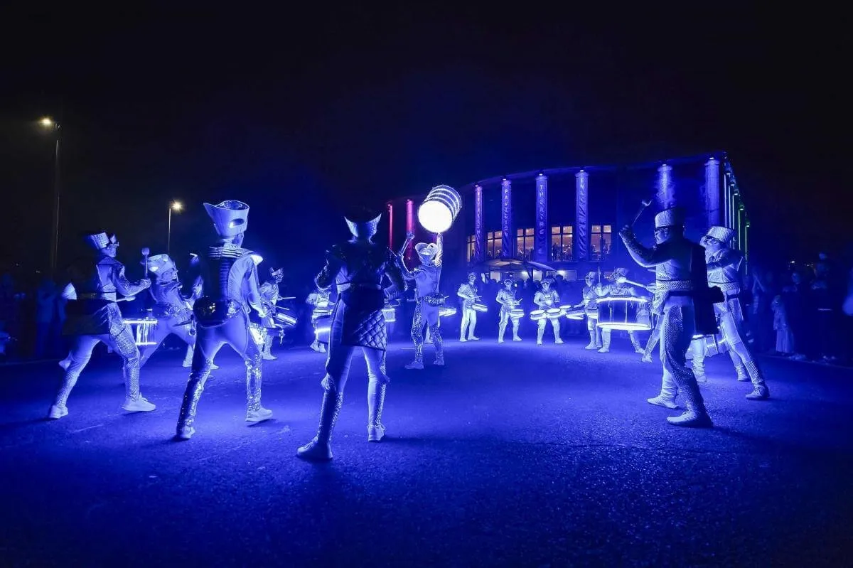 The combined Spark! and Fireflies ensemble circled together at night, drums glowing blue, a crowd gathered by a lit venue.