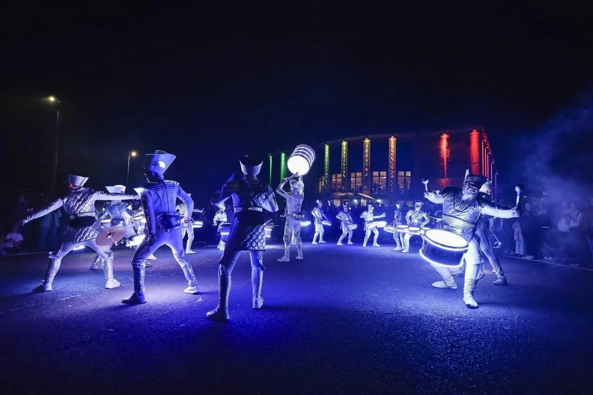 Fireflies performers in illuminated costumes drumming together in a wide blue-lit night scene.
