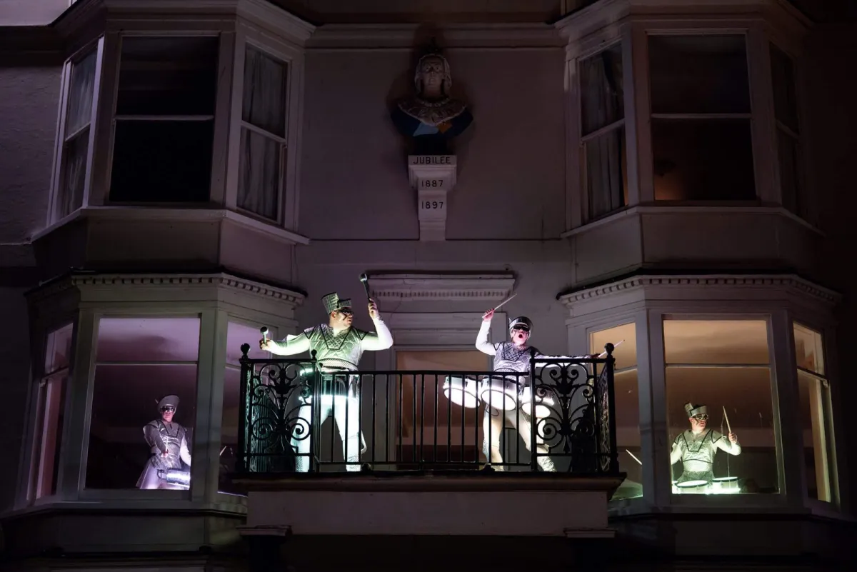 Fireflies performers drumming from the lit windows and balcony of a seafront house at night.