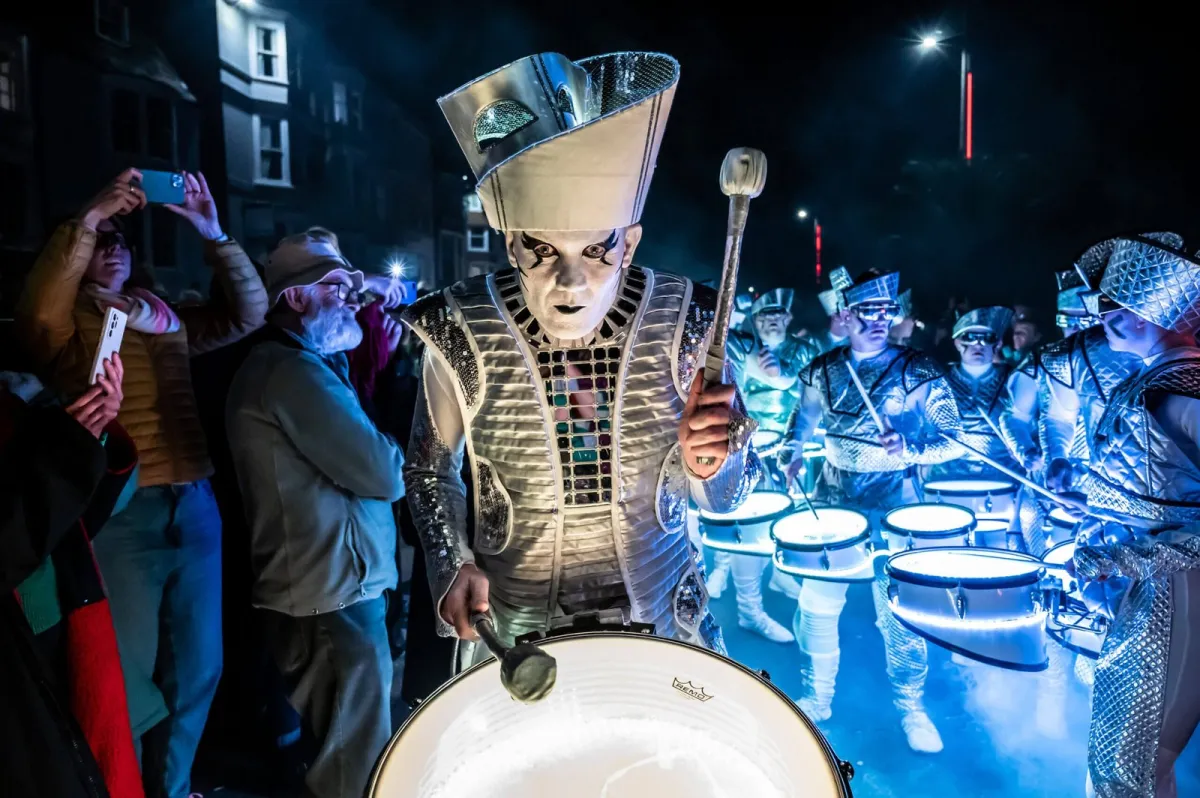 A Spark! performer leading the combined Fireflies ensemble past a crowd filming on their phones.