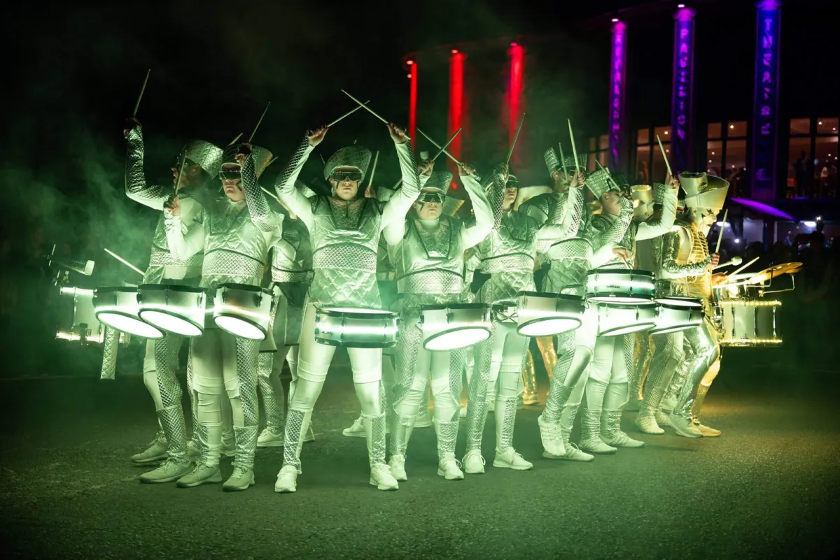 A large group of Fireflies performers in glowing white costumes, drumsticks raised together in the dark.