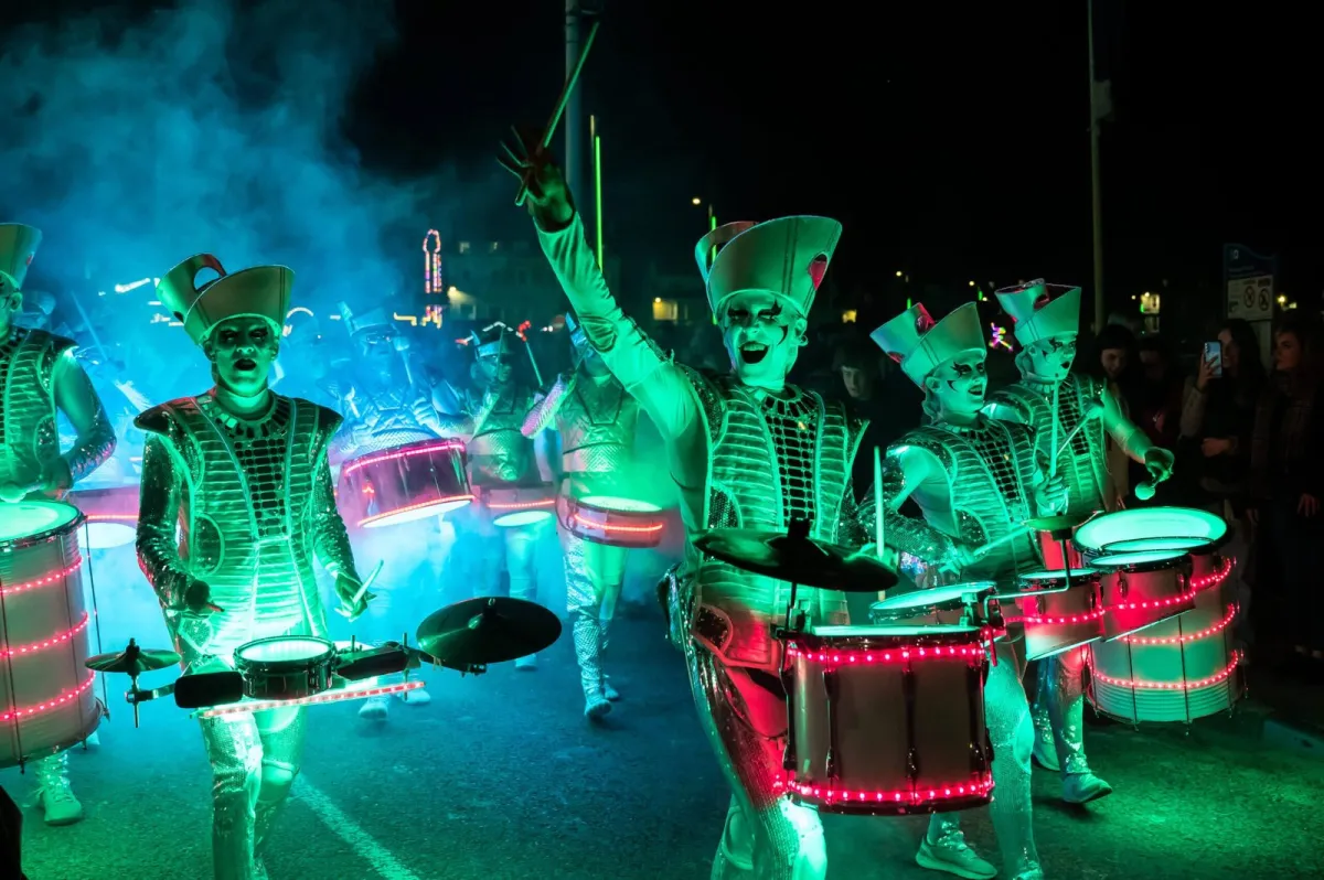 Fireflies performers in illuminated costumes parading at night, lit green and pink, arms raised.