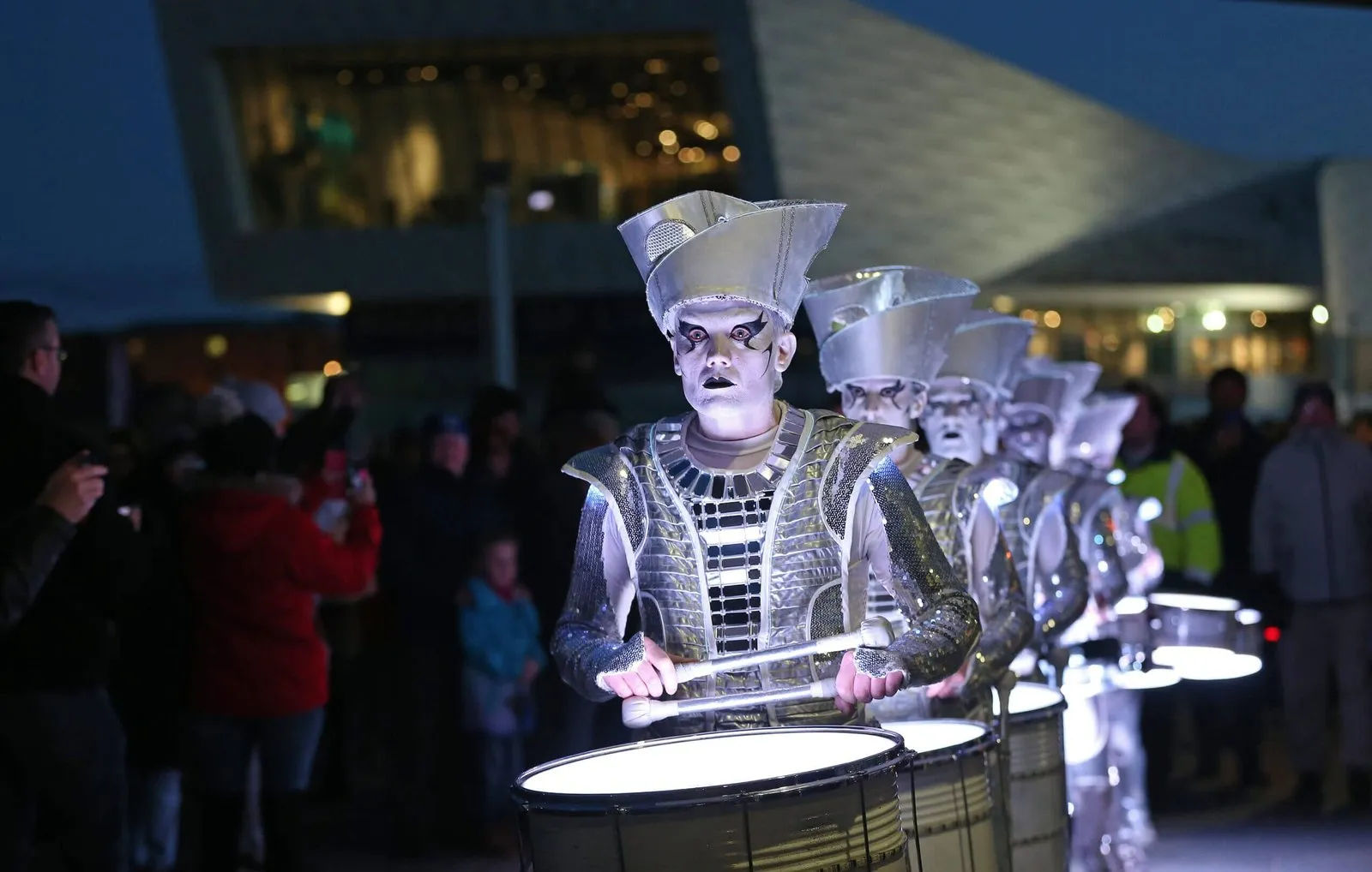 A line of Spark! performers drumming at dusk on the Liverpool waterfront.