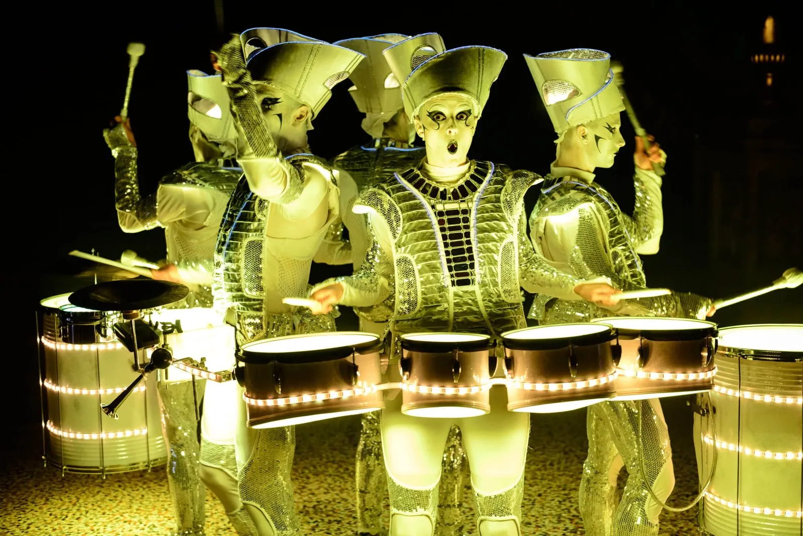 Spark! performers in a tight ensemble, illuminated drums glowing gold, mid-performance.