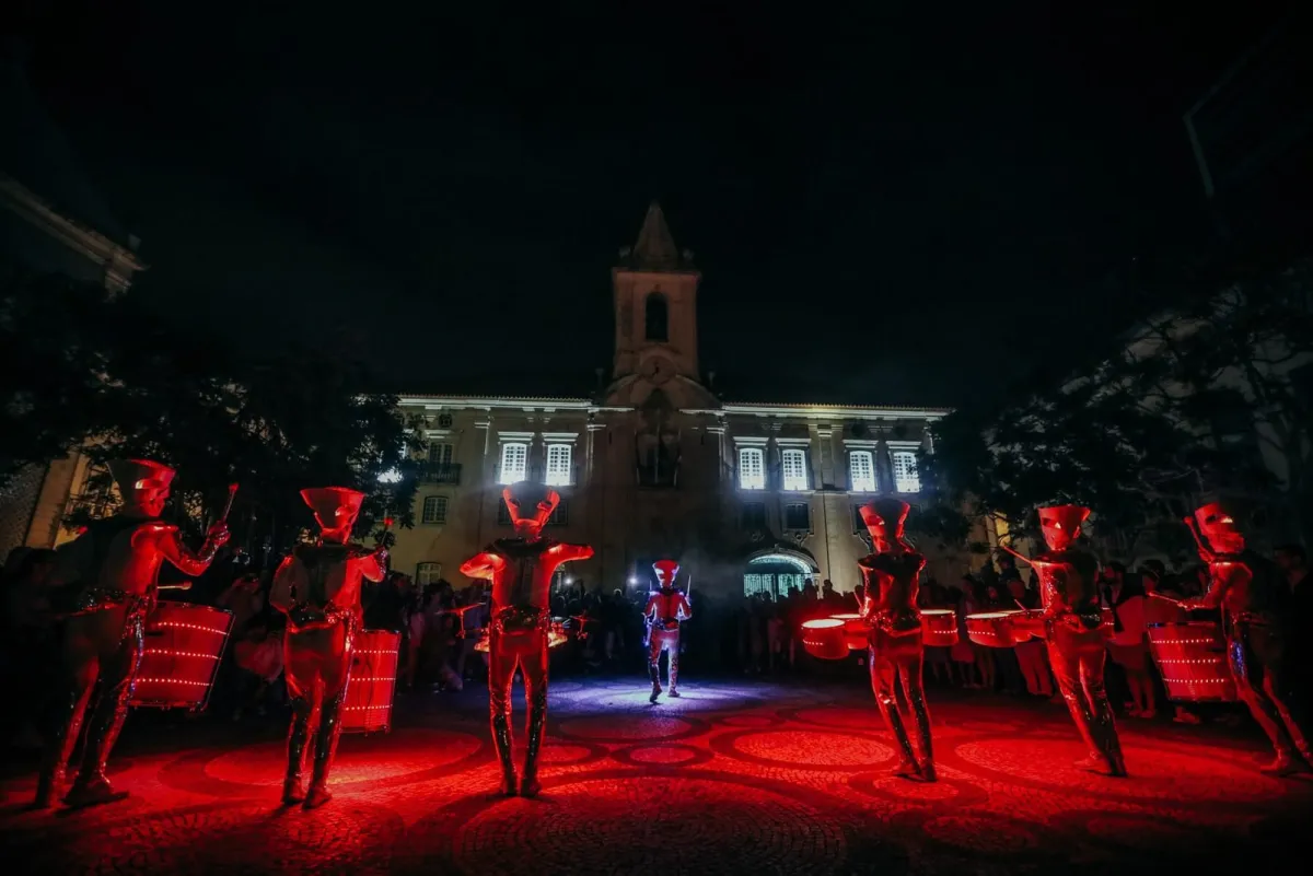 Spark! performers drumming in deep red light in a historic town square at night, crowd gathered.