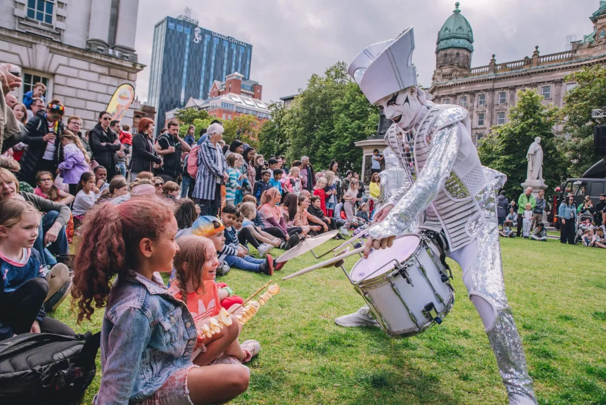 Spark! performers in silver leading a daytime city parade past Belfast City Hall.