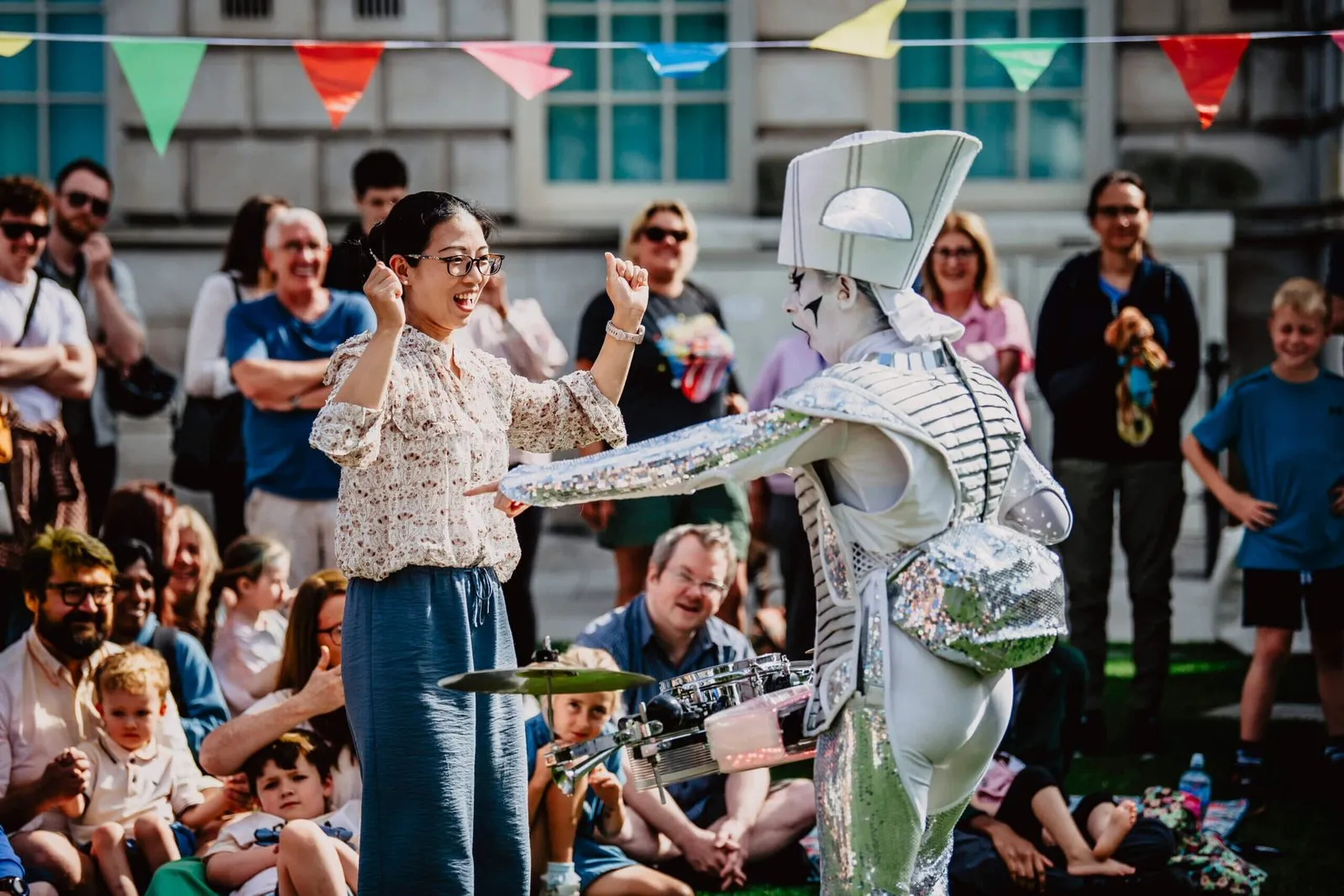 A Spark! performer in silver costume dancing with a delighted audience member at a daytime festival, crowd watching under bunting.