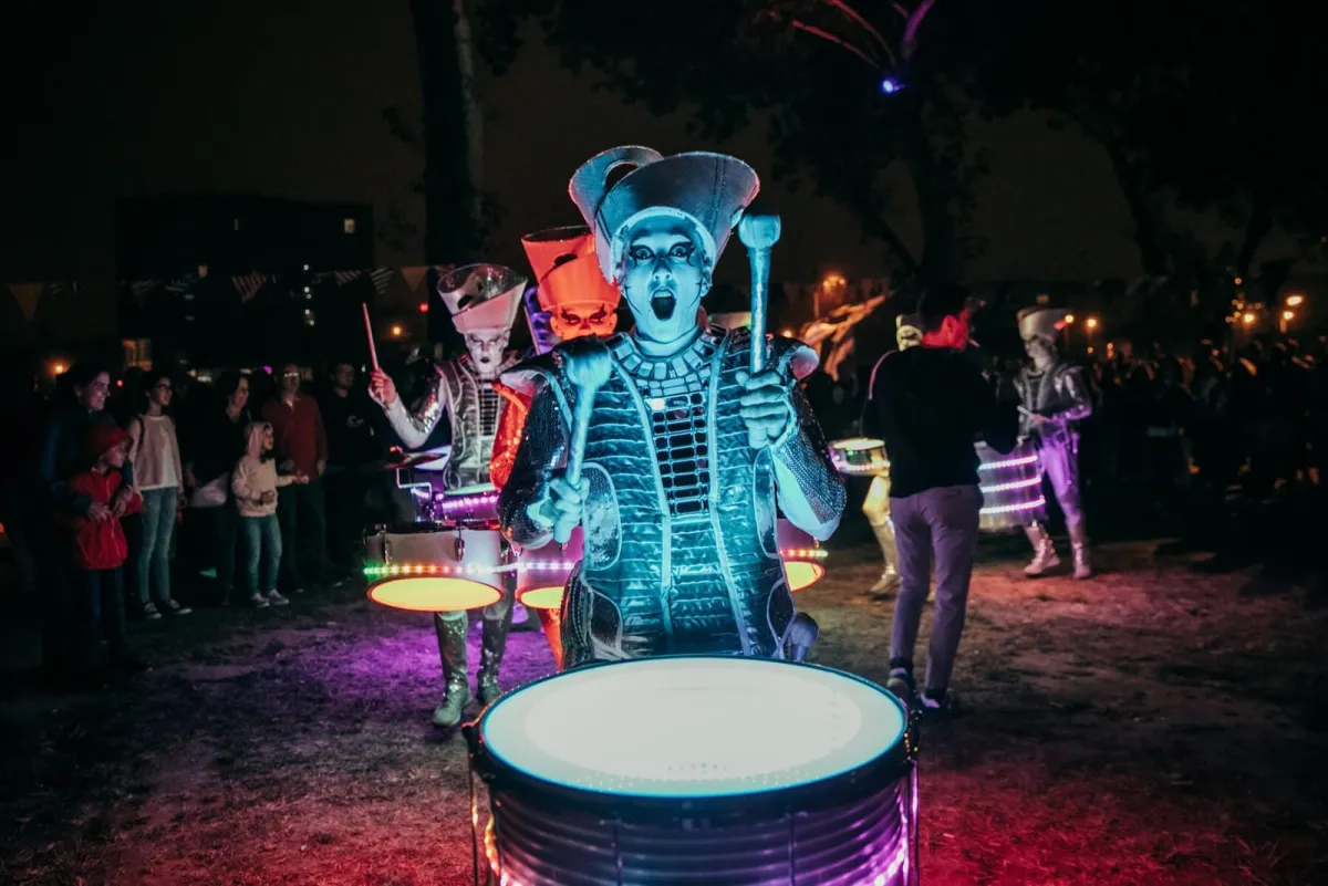 A Spark! performer on an illuminated barrel drum leading a night parade through a festival crowd.