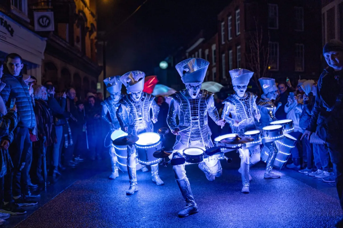 Spark! parading down a crowd-lined street at night, drums glowing electric blue.