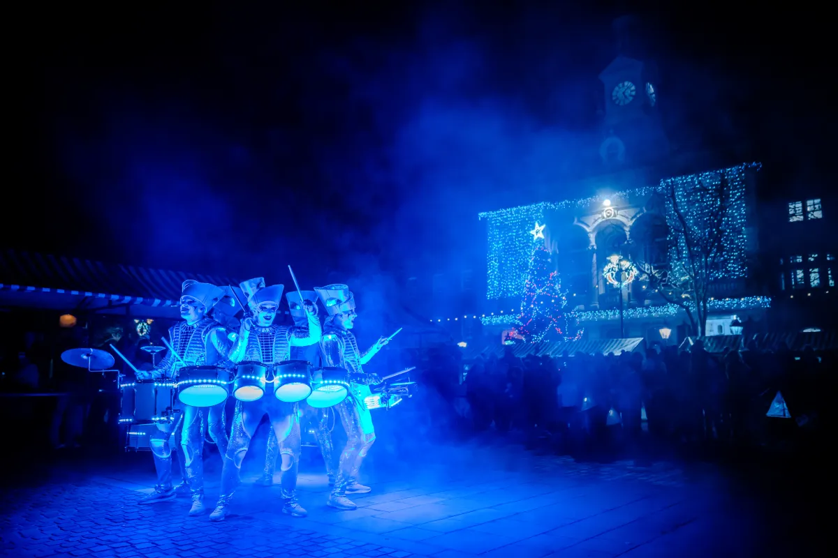 The Spark! cast drumming close together at night, costumes and drums glowing through blue smoke.