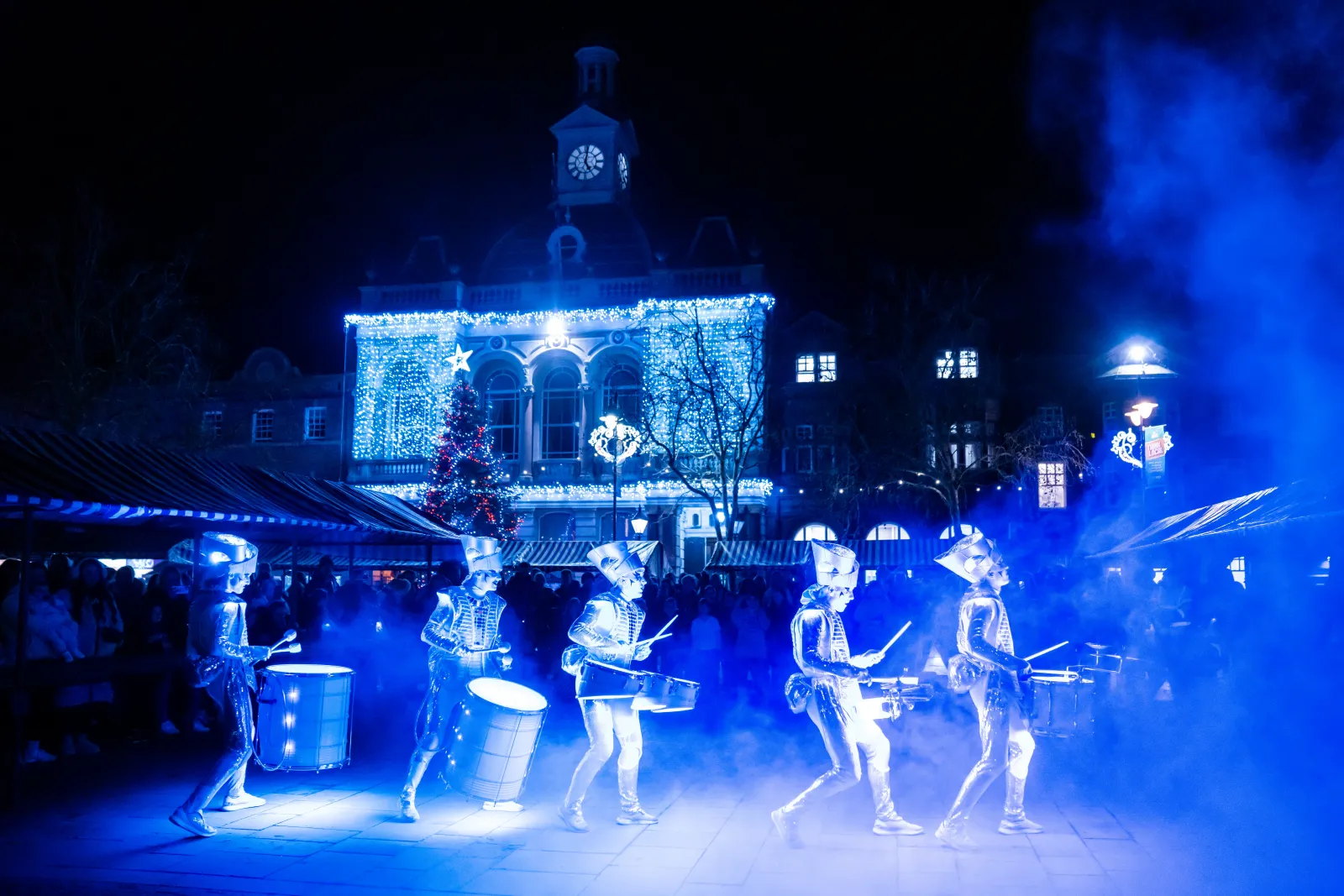 The Spark! ensemble drumming in blue light across a town square, festive town hall lit behind.