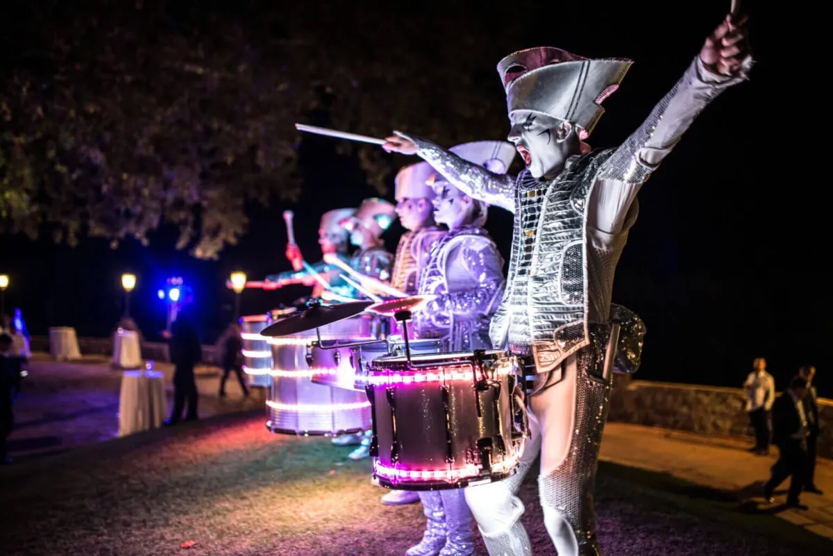 A Spark! performer mid-cheer with an arm raised, illuminated drums glowing pink at a night garden party.