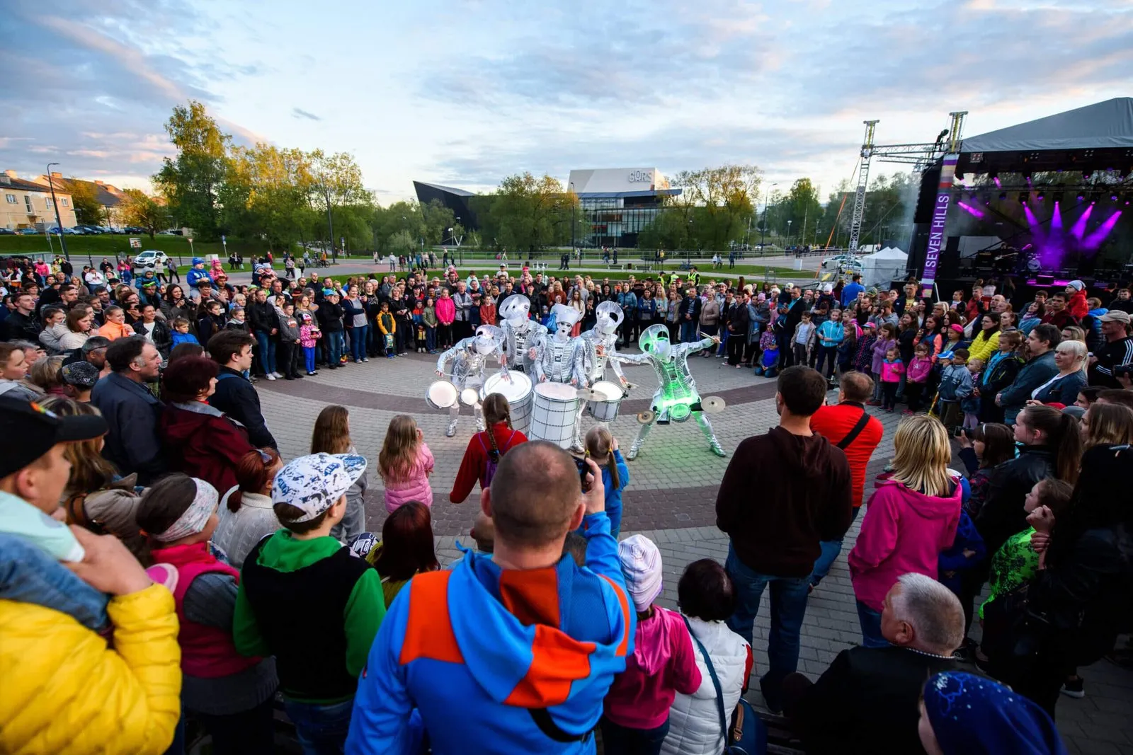 A huge dusk crowd circled around Spark! performers drumming in a park, stage lights glowing behind.