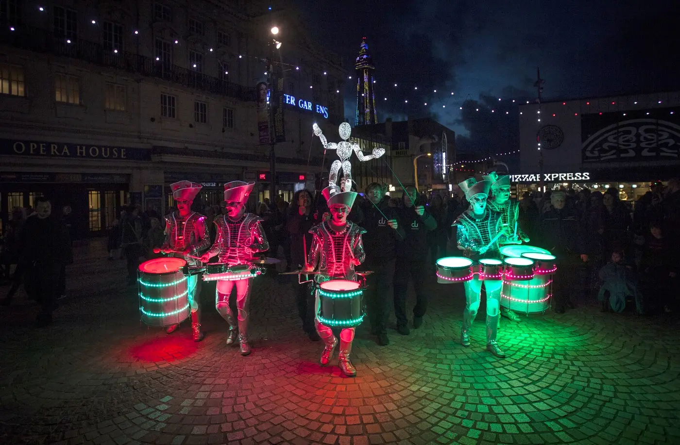 Baby Dundu riding above the Spark! drummers, lit red and green, in front of Blackpool's Winter Gardens.