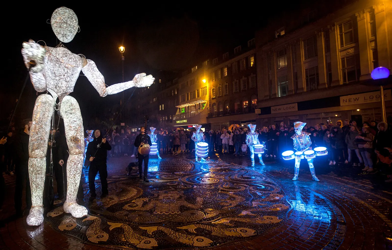 Dundu reaching out over a Bournemouth square at night while the Spark! drummers play on illuminated drums below.