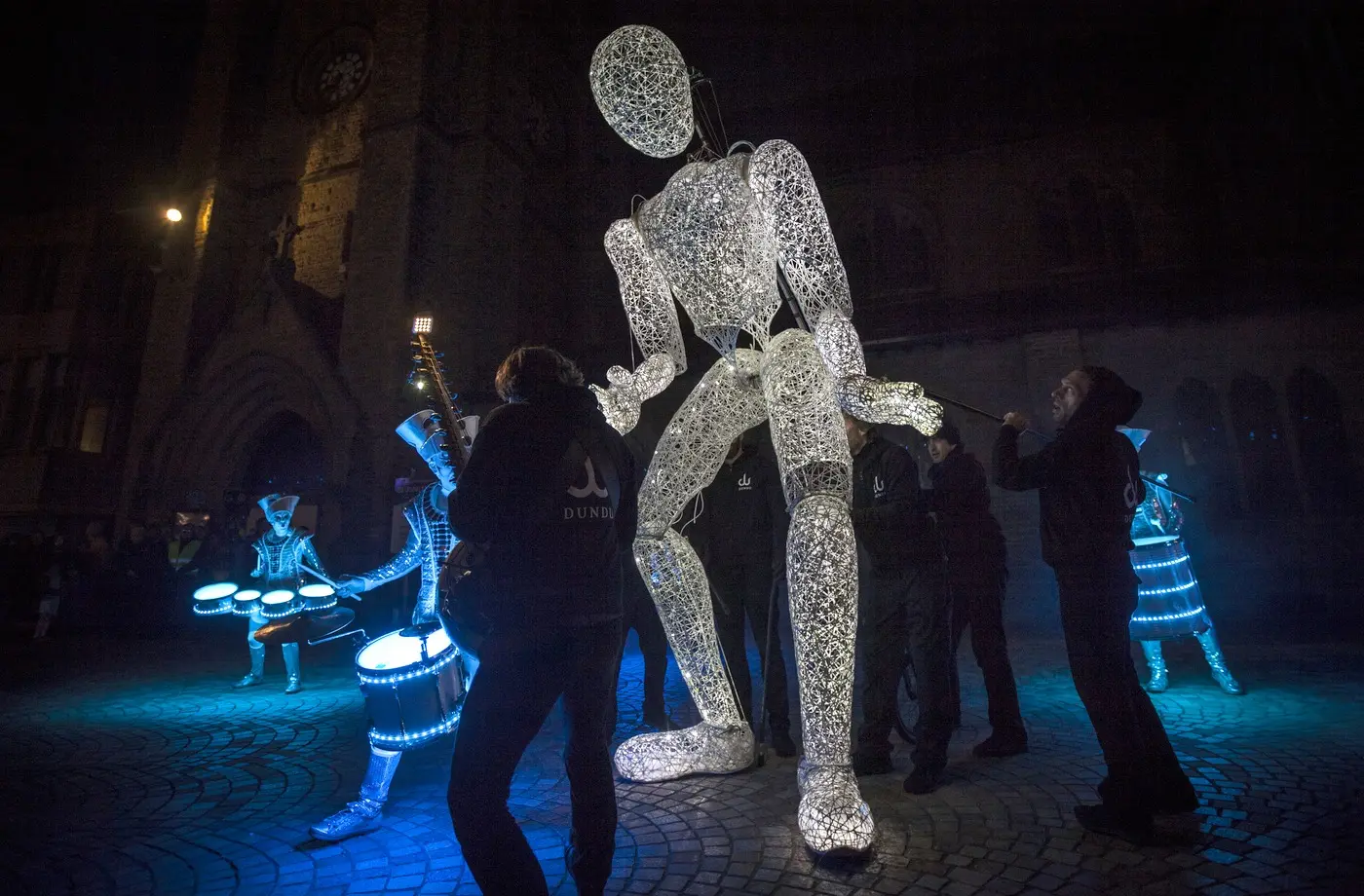 Dundu striding through the crowd at night with drummers lit blue on either side in Blackpool.