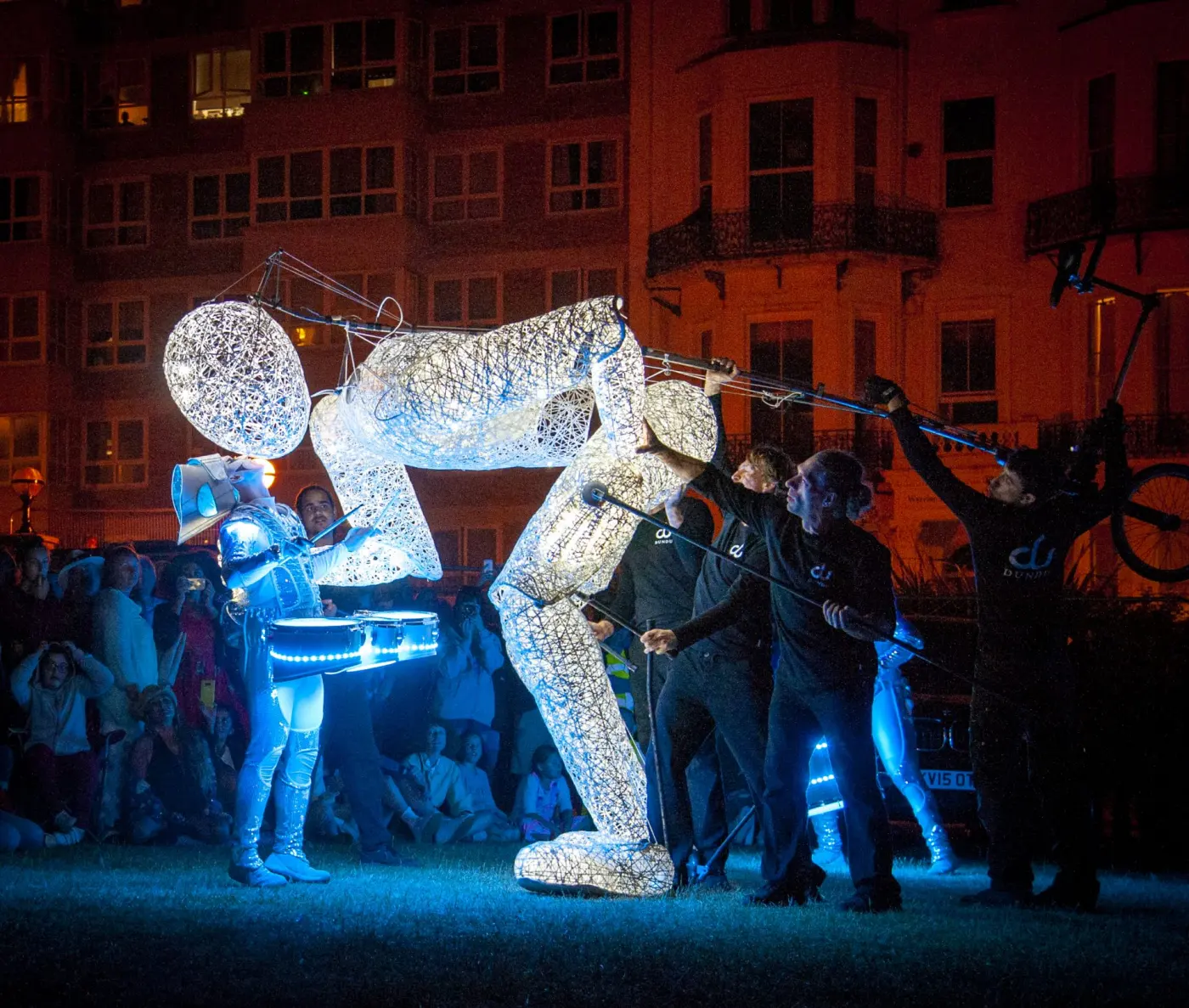 Dundu bending low to touch faces with a Spark! drummer, the puppeteers holding the giant's rods, in front of a red-lit seafront terrace.