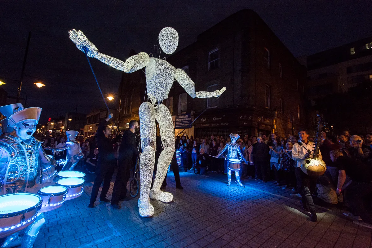 Dundu, a giant illuminated puppet, dancing through a packed street at night beside Spark! drummers on glowing blue drums.