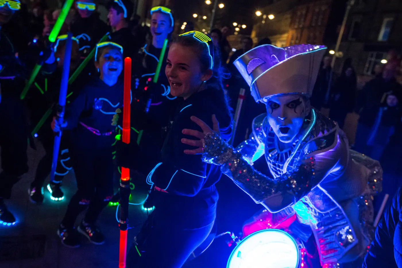 Children with glow sticks dancing and laughing beside a Spark! drummer in the Oldham crowd.