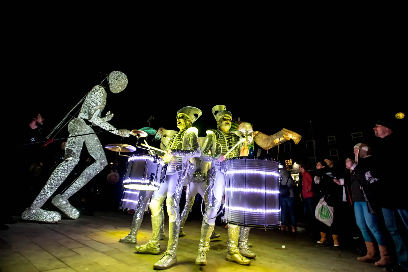 Dundu leaning in over Spark! drummers lit gold and green, the crowd close around them in Oldham.