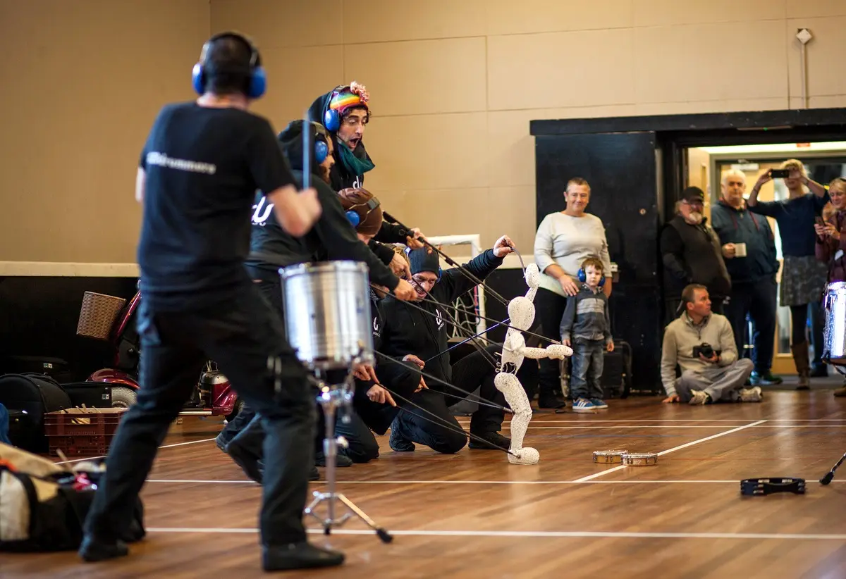 A Spark! drummer leading a community drumming workshop in a hall while Baby Dundu dances and families watch.