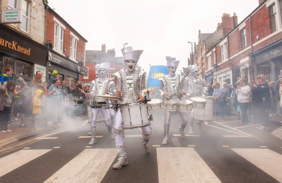 Spark! performers in white and silver parading up a shopping street in daylight, crowds either side.