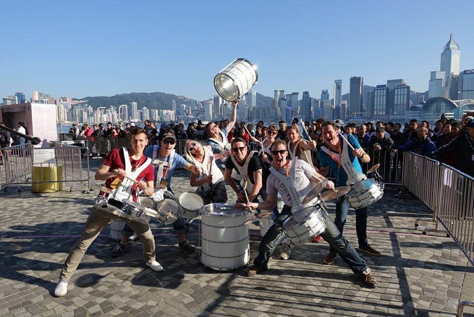 The Spark! team posing with their drums in front of a harbour city skyline on tour